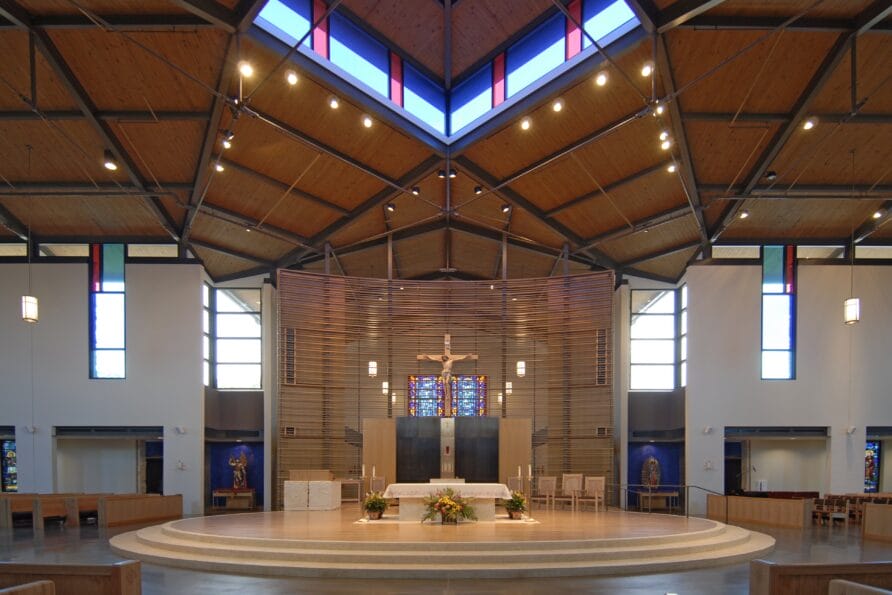 Modern church interior with crucifix, wooden ceiling, large windows, and altar surrounded by flowers.