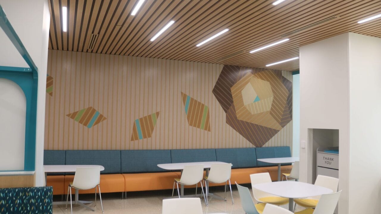 Modern cafeteria with abstract wall art, wooden ceiling, and white tables with colorful chairs.