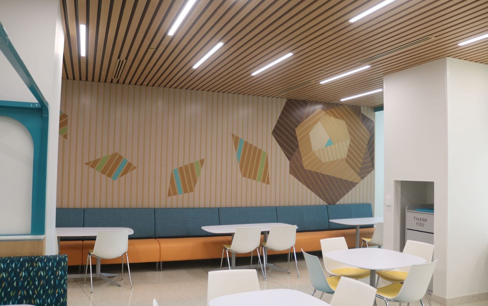 Modern cafeteria with abstract wall art, wooden ceiling, and white tables with colorful chairs.