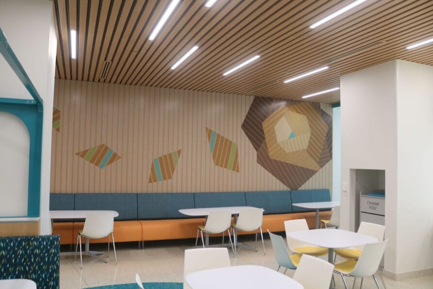 Modern cafeteria with abstract wall art, wooden ceiling, and white tables with colorful chairs.