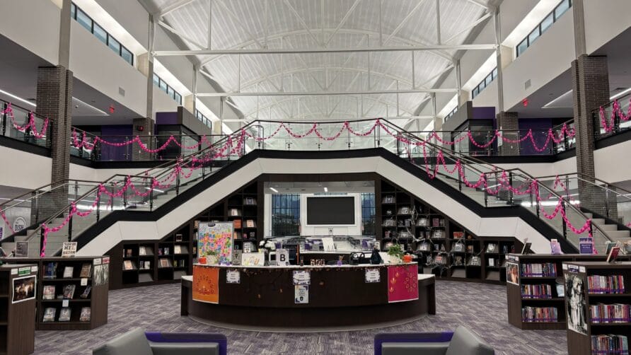 Modern library interior with central staircase, bookshelves, and seating area under high ceiling.