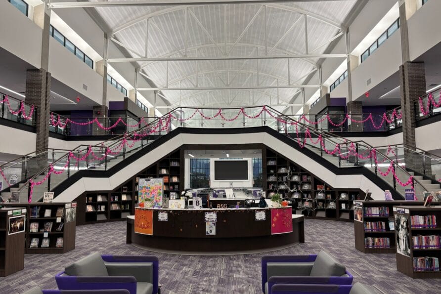 Modern library interior with central staircase, bookshelves, and seating area under high ceiling.