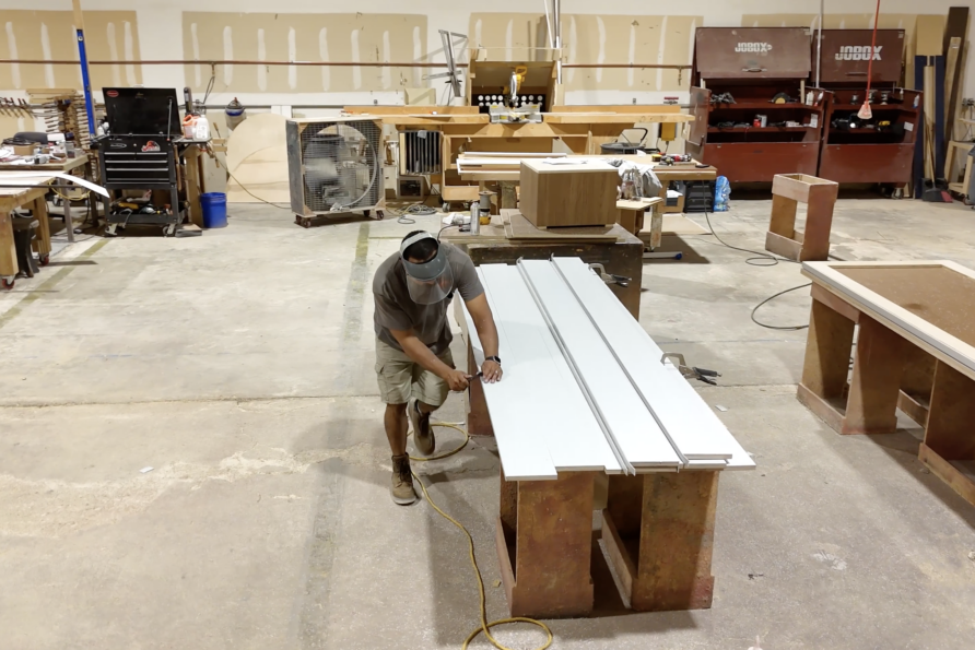 Worker sanding a long white wood plank in a spacious workshop with tools and machinery in the background.