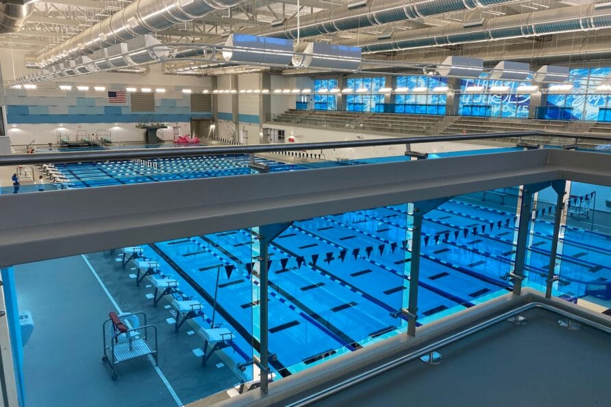 Indoor swimming pool with multiple lanes, bright overhead lights, and seating areas visible from an elevated view.