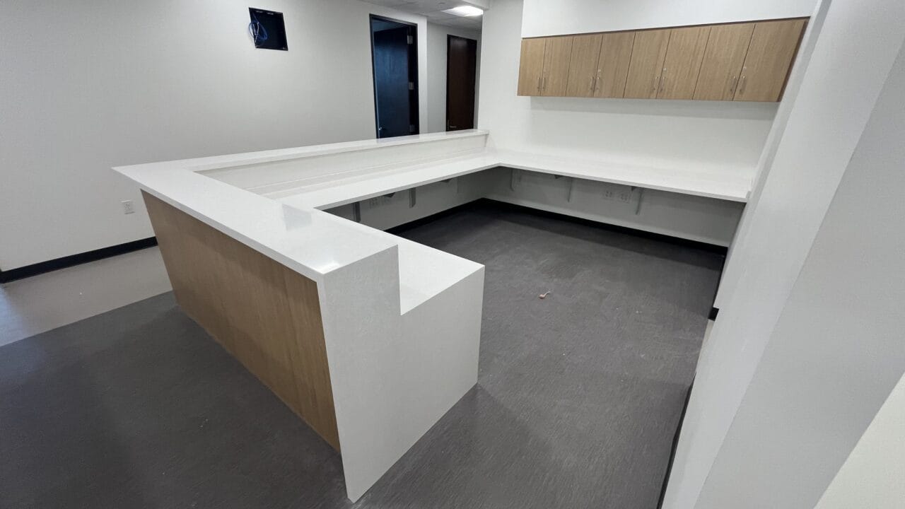 Modern L-shaped office reception desk with white countertop and wooden cabinets in a bright room.