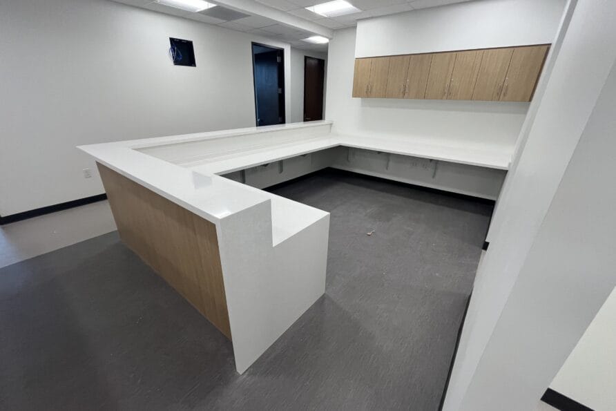 Modern L-shaped office reception desk with white countertop and wooden cabinets in a bright room.