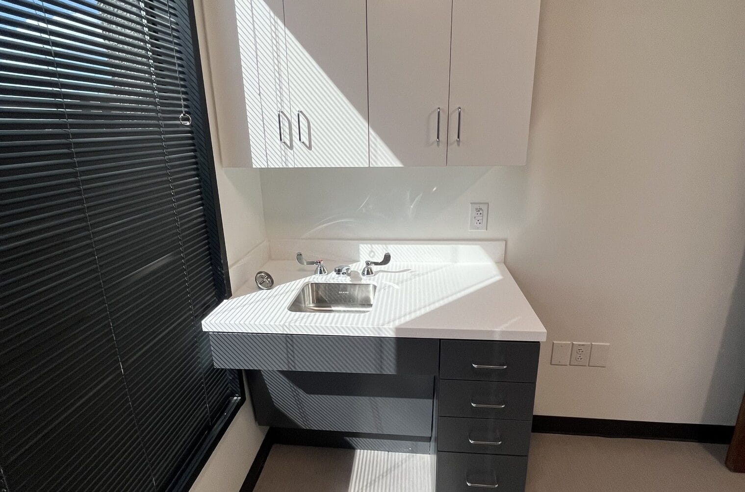Dental office sink with white countertop, under-cabinet storage, and natural light from a large window with blinds.